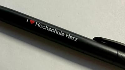 Detailaufnahme eines schwarzen Kugelschreibers mit dem Aufdruck I love Hochschule Harz auf einem weissen Blatt Papier