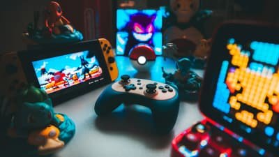 Spielkonsole, Controller und leuchtende Retro-Displays mit Sammelfiguren auf einem abgedunkelten Schreibtisch.