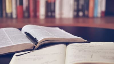Ein aufgeschlagenes Buch mit Stift und ein handschriftlich beschriebenes Notizheft vor einem Bücherregal